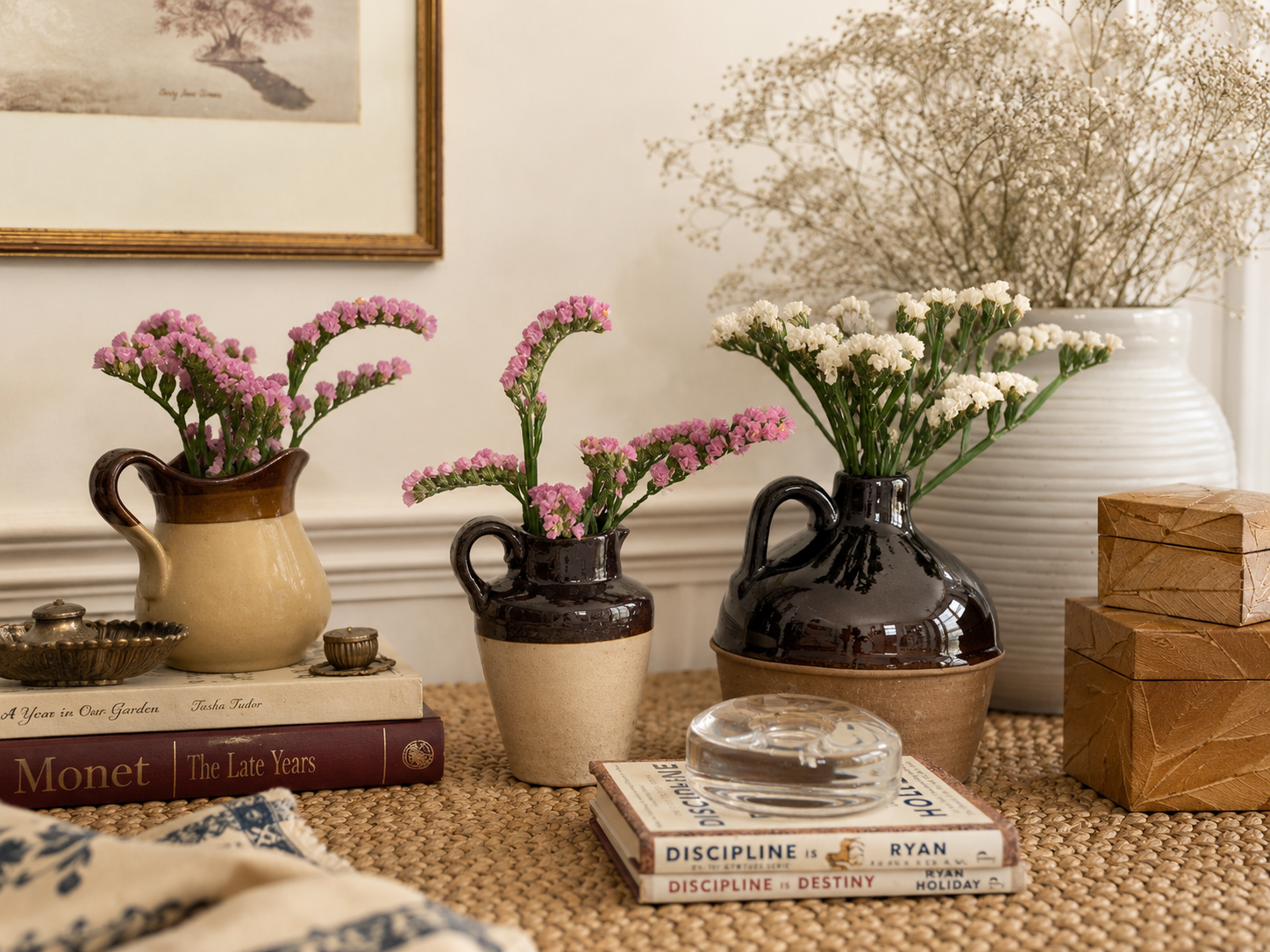 A warm collected home vignette with flowers, books, ceramic vessels, crystal, and meaningful objects styled on a woven table.